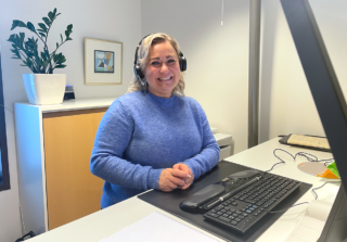 Asiakaspalvelun asiantuntija Sanna seisoo työpöytänsä ääressä kuulokkeet korvilla ja hymyilee.