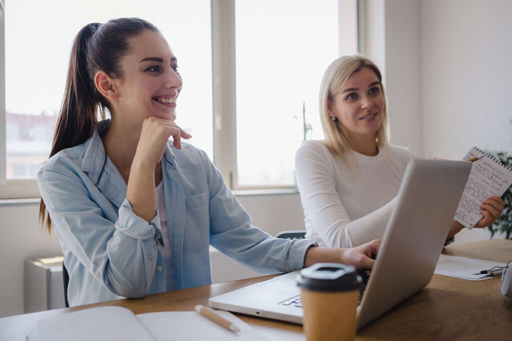 Kaksi naista istuvat pöydän takana ja hymyilevät. Pöydällä on tietokone ja pahvinen kahvimuki.
